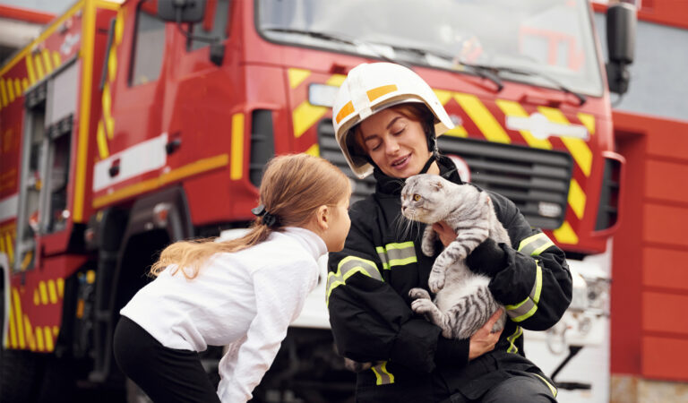 Firefighter kneeling in front of a fire truck, holding a rescued cat and comforting a young girl—highlighting pet safety during emergencies.