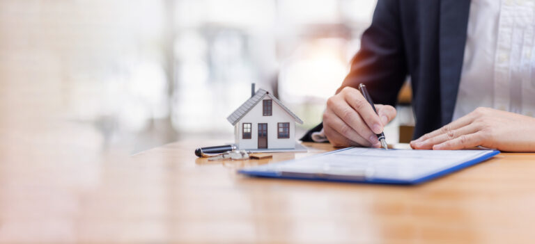 Person writing insurance notes on clipboard with house keys and model house during temporary housing review discussing additional living expenses